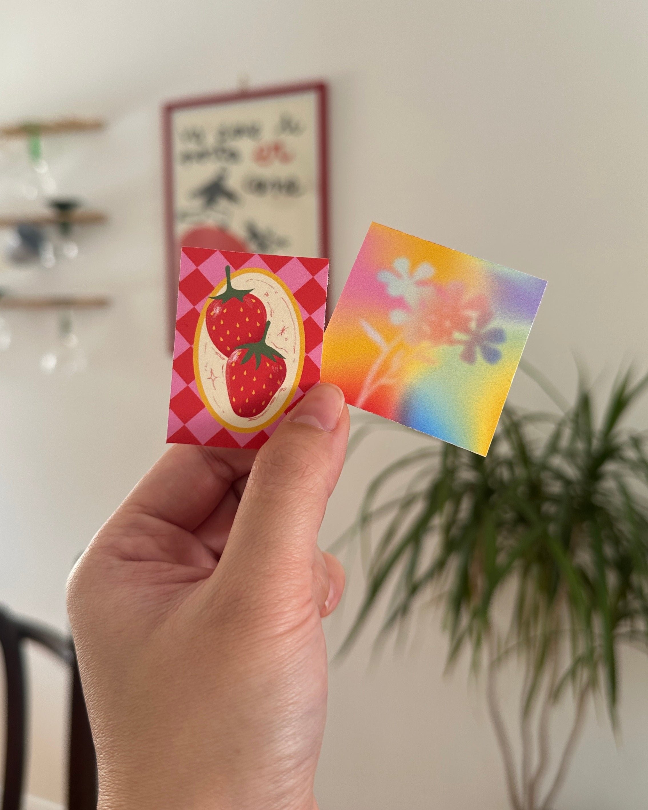 Hand holding two colorful cards with a plant and wall decorations in the background